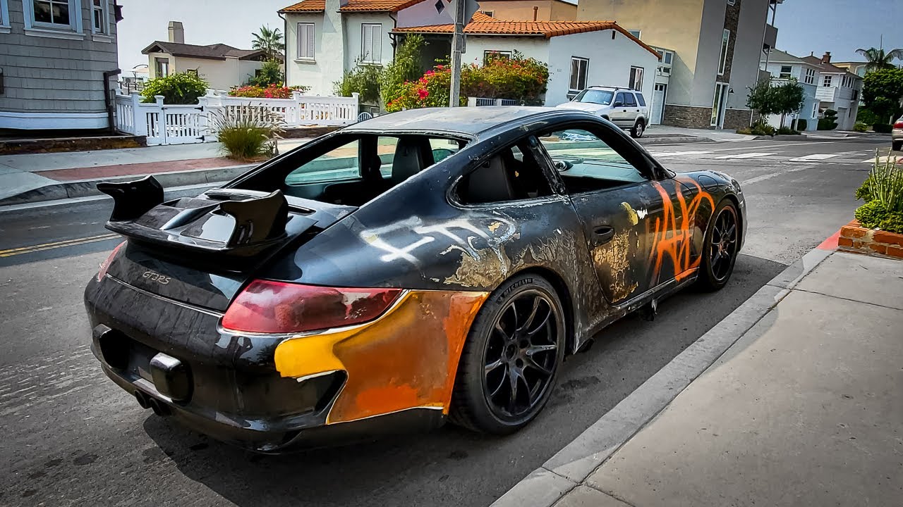 The 2007 Porsche 997.1 GT3 RS photographed in daylight on a Los Angeles residential street after recovery, showing spray-painted tags, missing rear wing, and fire damage to the rear quarter panel.