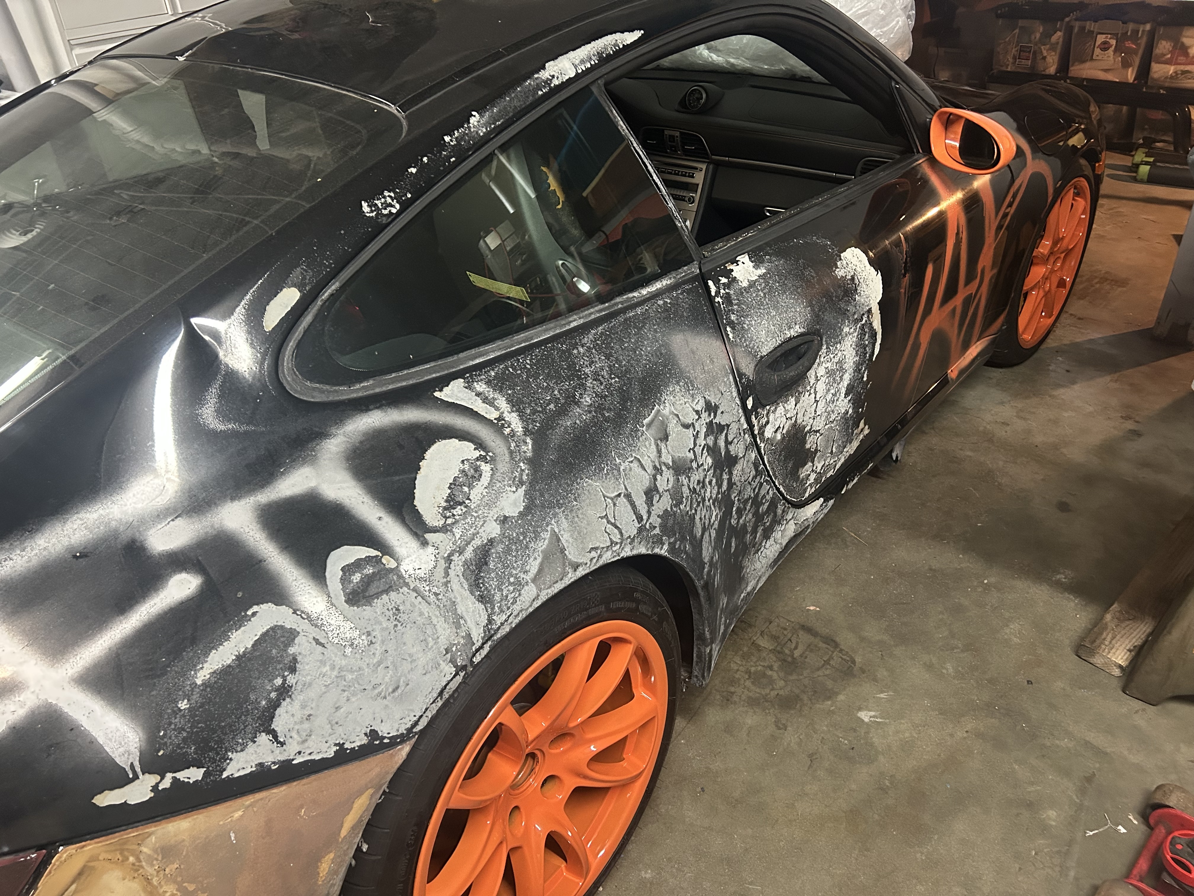 Close-up detail of the passenger-side door and rear quarter, showing burned and bubbled paint, areas of bare metal, residue from fire suppression, and orange spray-painted lettering across the bodywork. Wheels are factory-spec orange, restored to OEM by the current owner.