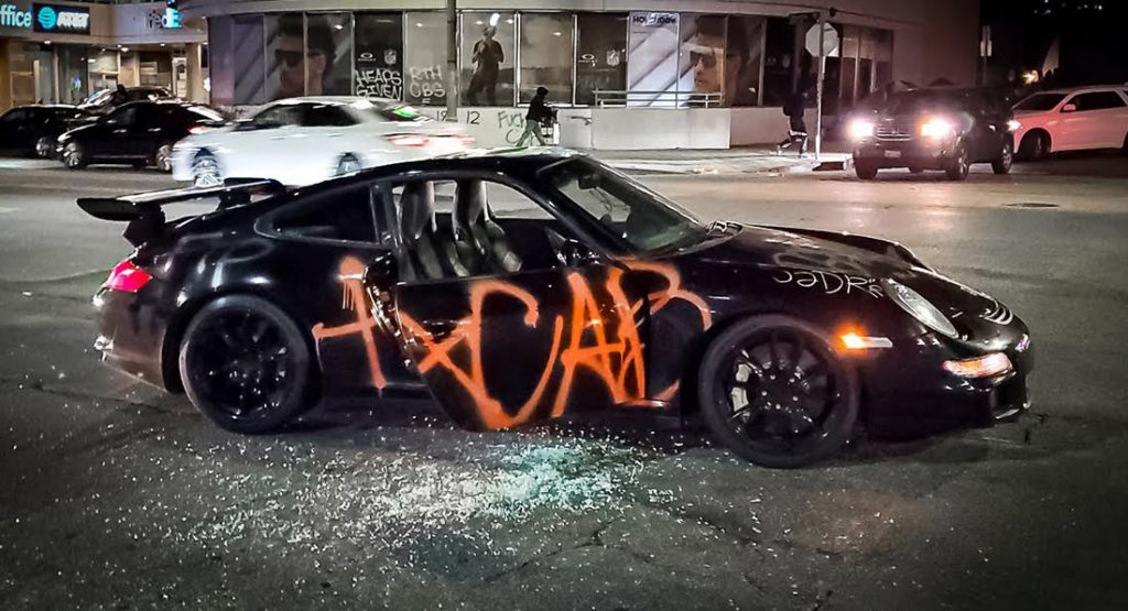 Archival photograph from the night of May 30, 2020 in Los Angeles. The Porsche is stopped in an intersection at night, driver door open, spray-painted with the letters A C A B, broken glass on the pavement, vehicles and a cyclist in the background.